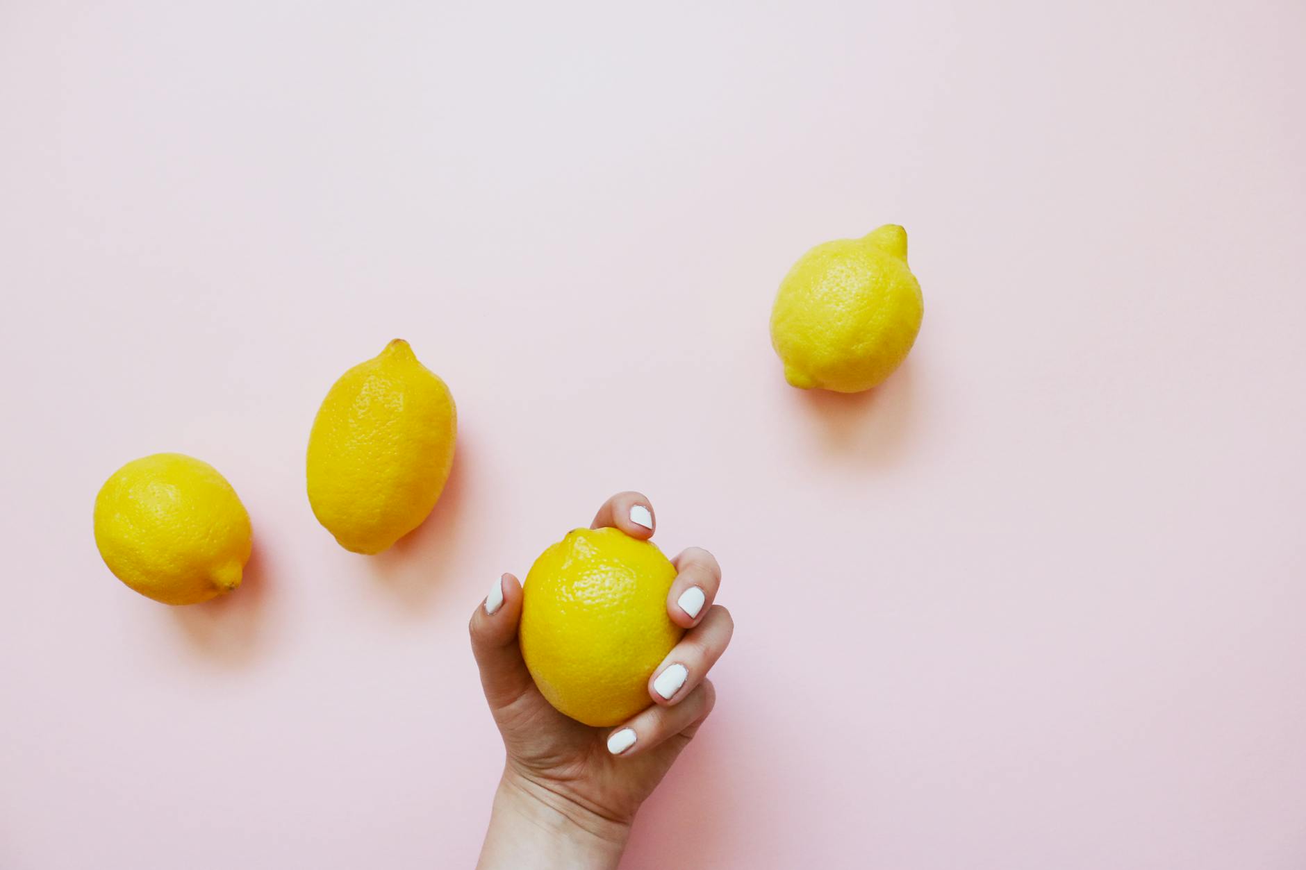 Hand holding a fresh lemon on a soft pink background surrounded by additional lemons