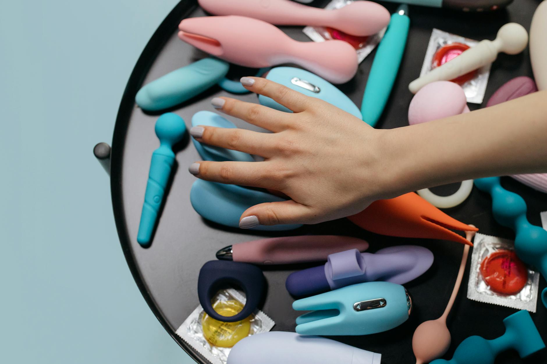 A hand reaching over a variety of colorful clitoral vibrators and wellness toys arranged on a light surface.